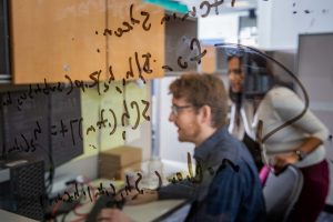 A glass pane with some equations written on it and two people working behind the door.