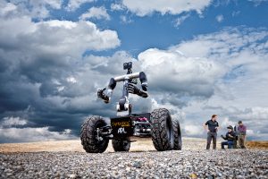 A man interacting with a huge Robot using the Virtual Reality gear, while two men are beside him where one of them is working on a laptop, in an open field.