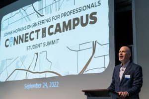A man wearing a blazer is speaking on a podium with a screen behind him displaying Connect to campus student summit poster