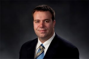 A headshot photograph of Jason Dever, wearing a suit, against a grey backdrop.