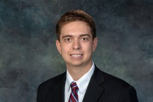 A professional headshot photo of Jordan Kremer, wearing a suit against a grey backdrop.