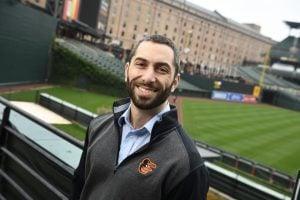Mike Synder smiles for a photo, wearing an Orioles pullover, standing beyond left field in Camden Yards. The field is behind him.