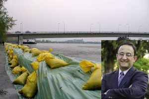 The edge of a river with yellow sandbags along the banks. A bridge is in the background. Inset is a photo of Gonzalo Pita, wearing a suit, with his arms crossed.