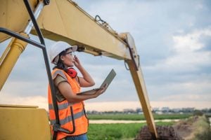 An environmental engineering holds a laptop in front of a backhoe digging into the earth.