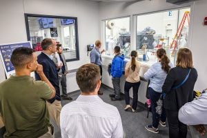 Students gathered in a room look through a window at the assembly of a space probe.