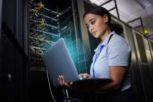 A woman works on a laptop connected to a server rack.