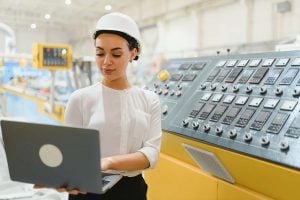 An engineer works on a laptop standing next to a large panel of switches in an industrial setting. 