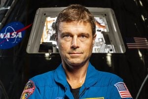 Portrait of Reid Wiseman in his flight suit in front of a NASA capsule. 