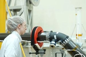 Person in a cleanroom suit and hairnet examines a large robotic arm or composite pipe assembly in a laboratory setting