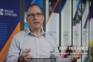 A man in a light blue button-up shirt is speaking in front of a backdrop of colorful banners. On-screen text identifies him as “BART PAULHAMUS, Chief, Intelligent Systems Center, Johns Hopkins APL,” and a subtitle at the bottom reads, “That’s computer science.”