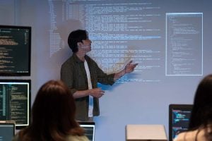 Presenter explains projected software code to a group of engineers using laptops during a technical team meeting.