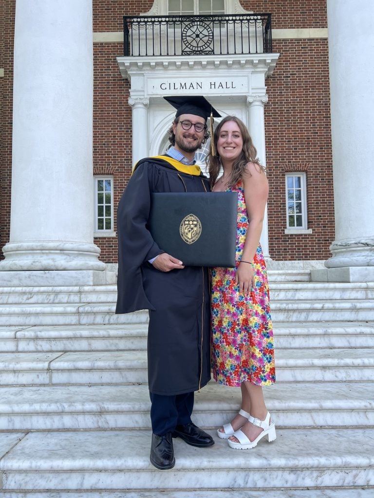 image of a man wearing a graduation cap and gown holding a diploma next to a woman where both of standing on steps in front of a building with the name "Gilman" in the background