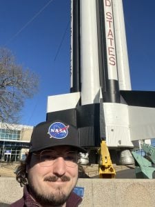 image of a man wearing a NASA hat standing in front of a rocket